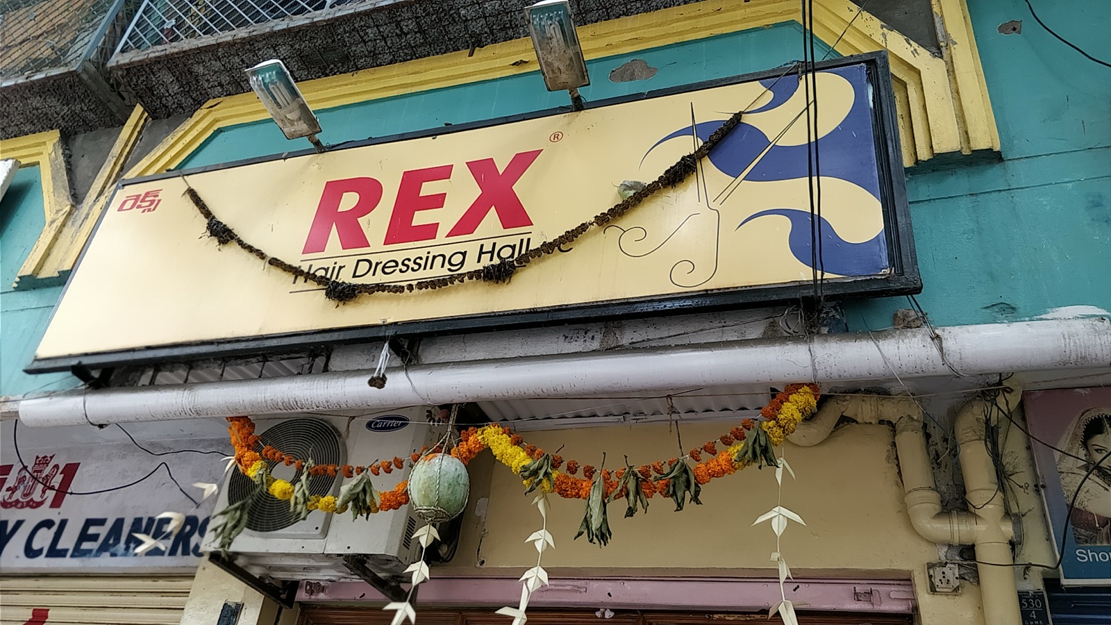 The hand-painted Rex Hair Dressing Hall street sign over the shopfront, garlanded with marigolds